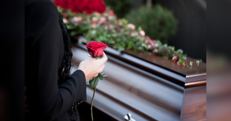 Caixão com flores por cima e uma pessoa de preto segurando rosa