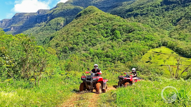 Duas pessoas em dois quadriciclos andando em uma estrada de chão cercada por vegetação e morros verdes. 