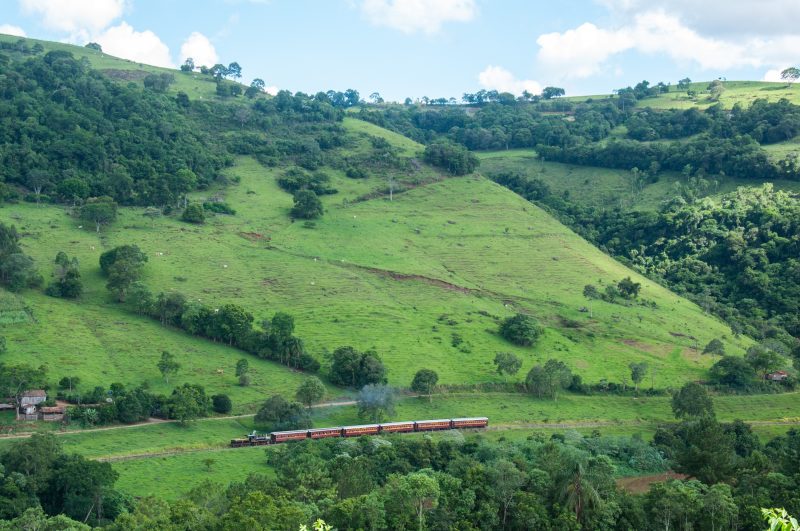 Foto aérea do trem Maria Fumaça com seis vagões de madeira passando ao lado de colinas verdes. 