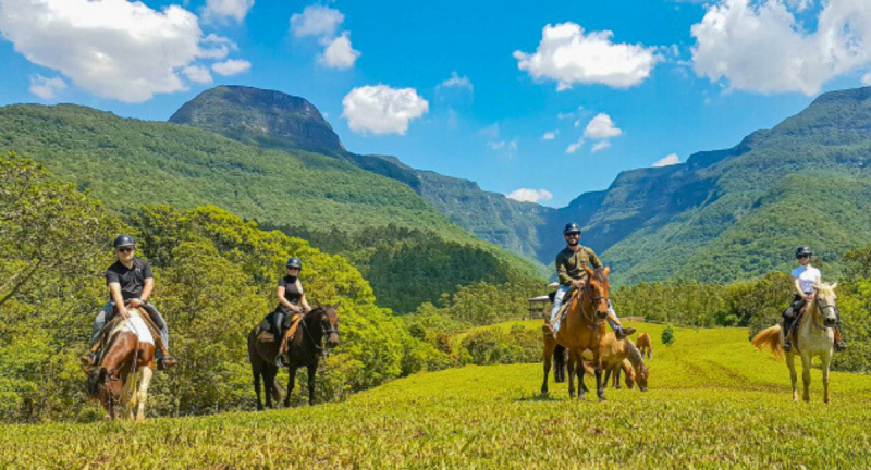 Passeio a cavalo Praia Grande Foto de quatro pessoas montadas em cavalos. Todas elas usam capacete e estão em um terreno gramado. Ao fundo, grandes paredões rochosos dos cânions de Praia Grande.