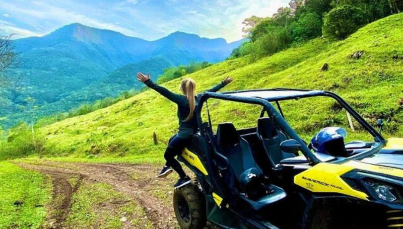 Passeio Off Road Praia Grande Foto de uma mulher jovem, loira e de cabelos compridos, de costas para a foto e com os braços abertos voltados para os cânions de Praia Grande. Ela está sentada na parte de trás de um UTV, veículo rústico com apenas dois lugares, aberto e com grandes rodas. O veículo está estacionado em uma estrada de terra com colinas verdes ao redor.