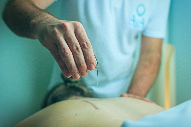 Foto mostra a mão de um homem despejando uma pequena quantidade de óleo nas costas de uma mulher deitada. Não aparece o rosto de nenhum deles. 