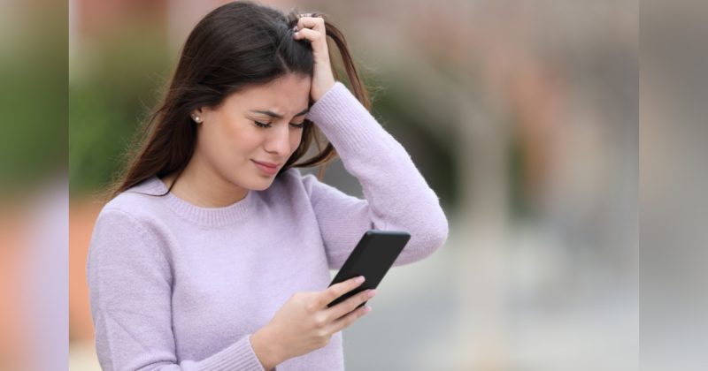 Pessoa de blusa lilás com celular na mão com cara de tristeza