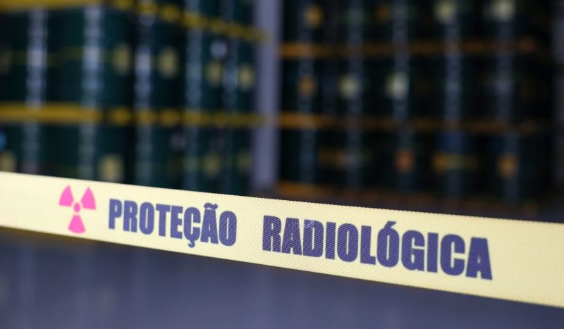 Faixa com a descrição de 'proteção radiológica' em primeiro plano e imagens de cilindros em desfoque ao fundo