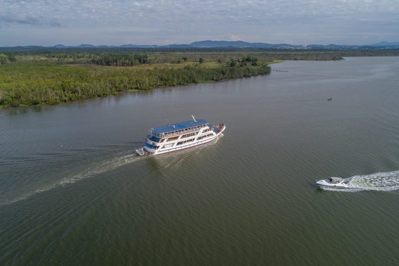 Foto aérea do Barco Príncipe, grande e nas cores branca e azul, navegando pela baía. Ao fundo, vegetação. 