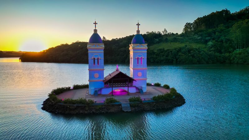 Torres brancas de uma igreja com uma luz rosa em janelas. Ao redor dela, uma área cimentada e cercada pelo lago do Rio Uruguai. Ao fundo, um morro verde e o sol se pondo atrás dele. 