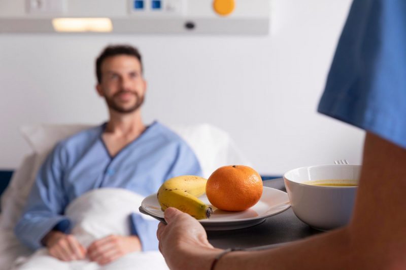 Homem desfocado na cama do hospital recebendo um prato com frutas e sopa 