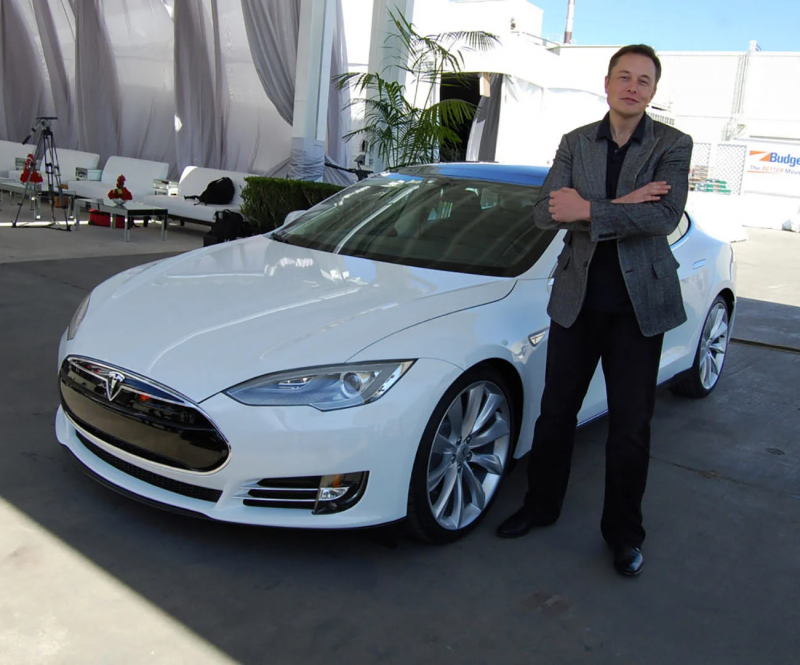 Elon Musk, vestido com roupas escuras, ao lado de um carro branco da Tesla