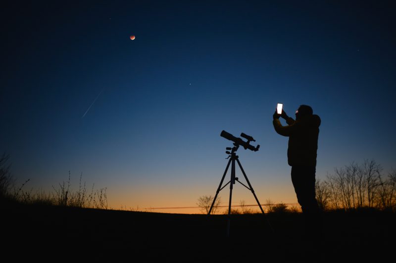 Astrônomo olhando para o céu estrelado com planetas, estrelas cadentes e eclipse lunar total, a Lua de sangue, com um telescópio enquanto usa o smartphone 