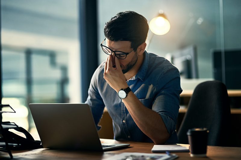 Homem com estresse, com a mão no rosto em frente ao laptop 