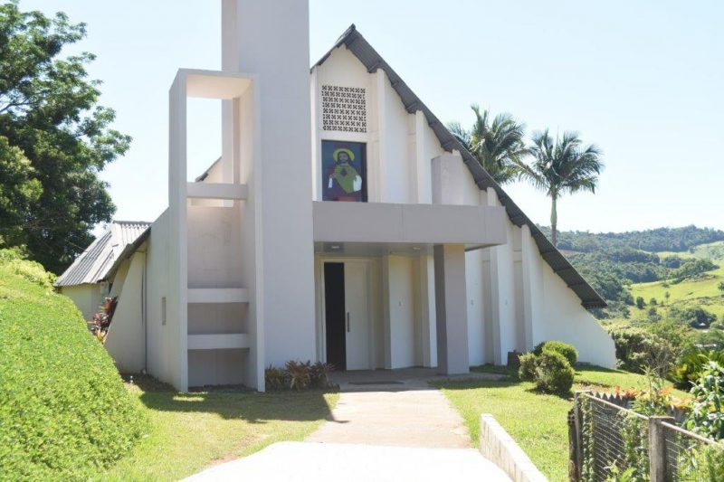 Igreja de paredes claras com telhado triangular, que vai quase até o chão. Na fachada há algumas colunas decorativas encostadas na parede. Em cima da porta, na parte superior da igreja, uma imagem de Jesus Cristo. Ao redor da igreja, colinas verdes.