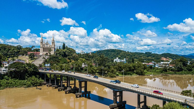 Foto aérea de Gaspar. Em primeiro plano, uma ponte de concreto que cruza um rio com águas barrentas. Sobre a ponte passam vários carros. Ao fundo, a vegetação que cerca o rio, alguns morros verdes e a Igreja Matriz de Gaspar, grande e com duas torres à vista. O dia está ensolarado e o céu azul, com algumas nuvens brancas. 