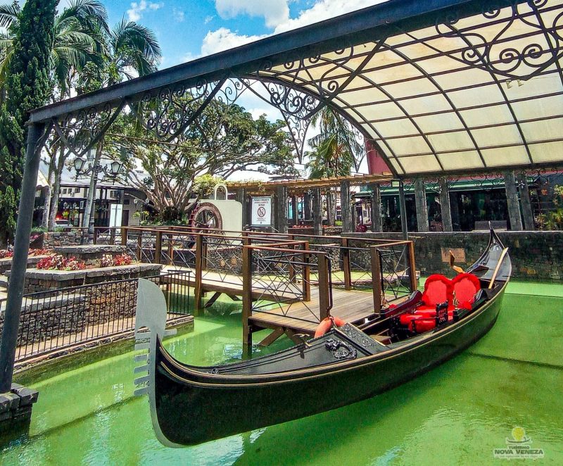 Foto de uma embarcação a remo comprida e estreita na cor preta. Dentro dela há dois assentos estofados na cor vermelha. Ela está em um lago artificial de água esverdeada e debaixo de uma estrutura de ferro preta com cobertura clara. Ao lado da embarcação há uma passarela de madeira por onde as pessoas chegam até ela. Aparece também algumas árvores e canteiros de flores da praça central. 