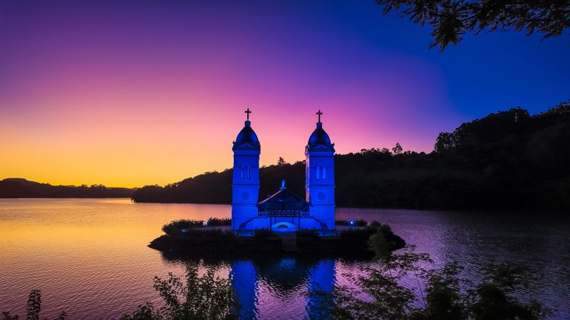 Duas torres de uma igreja instalada em uma espécie de ilha de concreto, cercada pelo lago do Rio Uruguai. Ao fundo, vários morros verdes. O céu está em cores de azul, amarelo e rosa, ao anoitecer. 