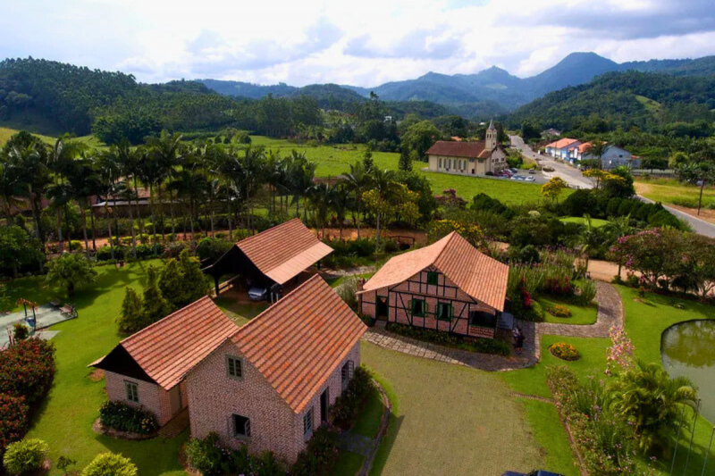 Vista aérea de algumas casas enxaimel, de tijolos à vista e ripas de madeira intercaladas nas paredes, instaladas em uma ampla área gramada, cercada por árvores e colinas. Ao fundo, aparece uma pequena igreja de torre única e uma estrada asfaltada. 