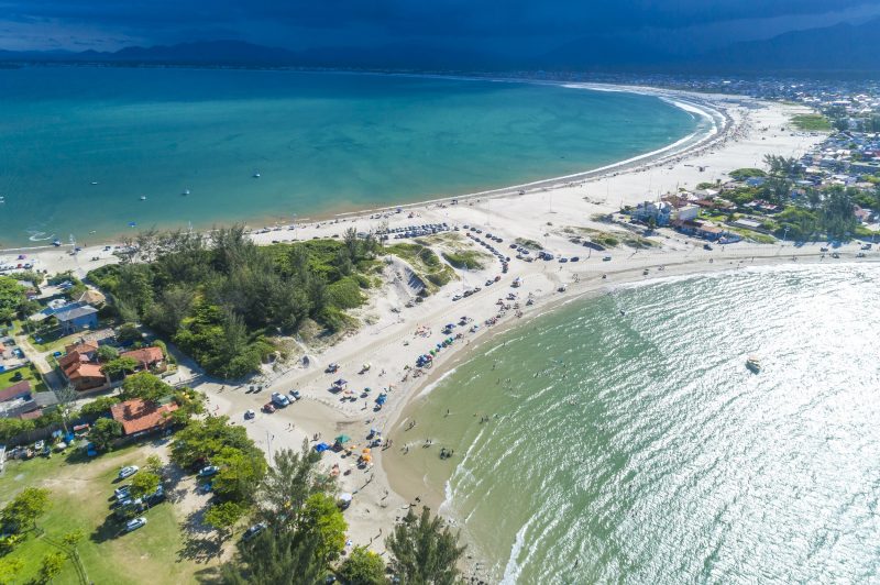 Foto aérea das praias do Sonho e da Ponta do Papagaio. Na parte inferior da foto, o mar cristalino da Praia do Sonho, onde estão algumas pessoas. No canto esquerdo, ao lado do mar, há uma área gramada com árvores e alguns carros estacionados. Na parte superior da foto, o mar da Ponta do Papagaio. Entre os dois mares, no meio da foto, há um trecho de faixa de areia que une as duas praias e as duas frentes para o mar. Nesse trecho, há carros estacionados, uma pequena área de vegetação e algumas casas. 