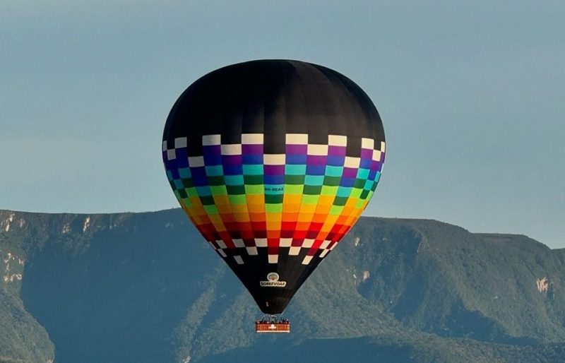 Balão colorido voando sobre grandes paredões rochosos. Ao fundo o céu azul. 