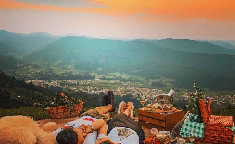 Na parte inferior da foto, um homem e uma mulher jovens e de pele clara estão deixados em uma área gramada alta. Eles se olham e sorriem. O rosto deles aparece parcialmente. Ao lado deles, há uma cesta de piquenique e um caixote de madeira, sobre o qual há taças e uma bebida dentro de um baldinho de inox. Ao fundo, aparecem morros verdes e o Centro de Urubici, com centenas de construções. O céu está em cores de azul e laranja, durante o pôr do sol. 