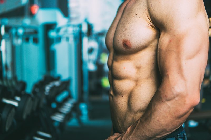 Homem de perfil, sem camisa na academia para mostrar ganho de massa muscular