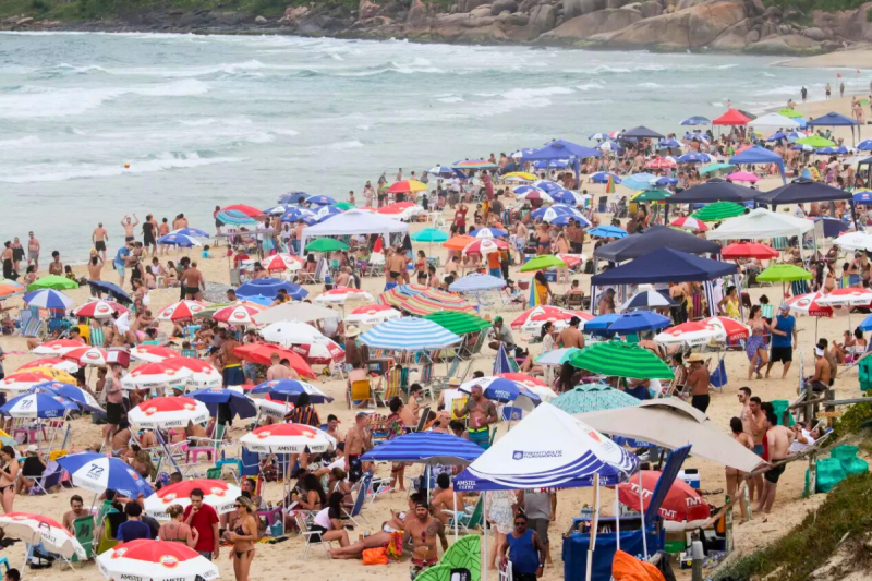 Foto aérea da areia da Praia Mole tomada por centenas de guarda-sóis coloridos e pessoas. Ao fundo, aparece uma parte do mar de águas claras com onda e um costão rochoso. 