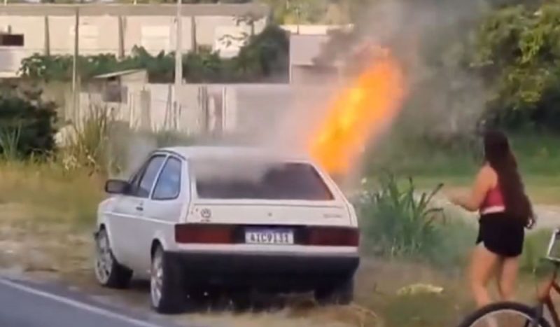 Mulher incendeia carro errado ao tentar se vingar do marido no Paraná. Na imagem, o veículo em chamas com a mulher ao lado.