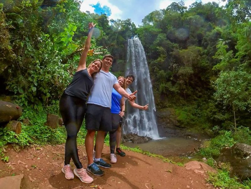 Foto de quatro pessoas jovens, dois homens e duas mulheres, em frente à uma cachoeira cercada pela vegetação em Praia Grande. Todos eles olham para a foto, sorriem e acenam.
