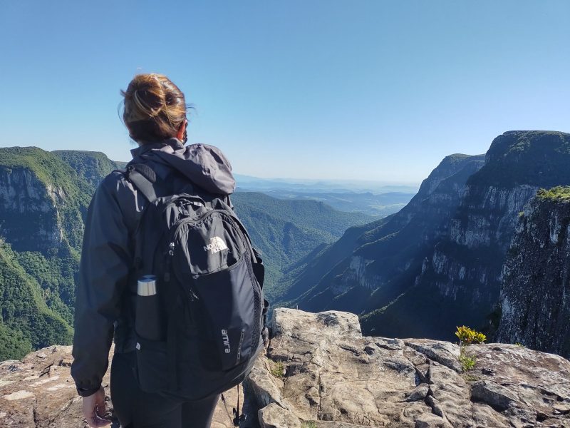 Cânion Fortaleza Mulher jovem, de cabelos castanhos e presos, usando uma japona e uma mochila preta, de costas para foto. Ela está no alto do Cânion Fortaleza e observa os grandes paredões rochosos à sua frente.