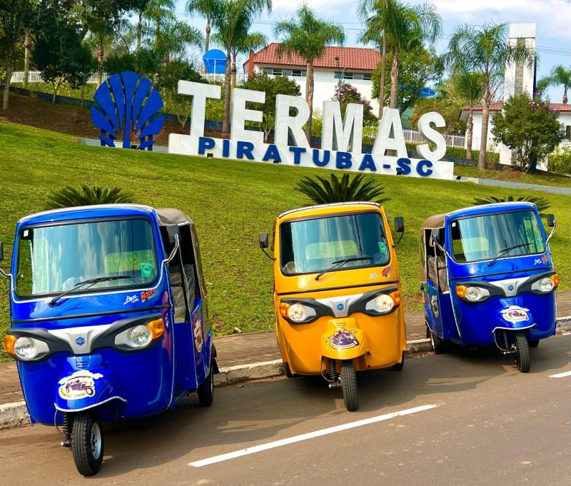 Foto de três veículos tuk-tuk, dois azuis e um amarelo. Eles são veículos pequenos, de três rodas e cobertos na parte de trás com uma espécie de tecido. Eles estão estacionados em uma rua e atrás há uma área gramada com um totem escrito "Termas Piratuba -SC". 