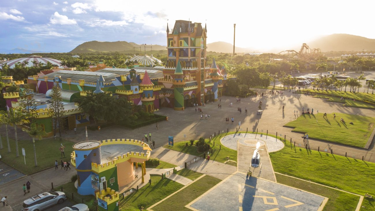 Foto aérea do castelo multicolorido do parque Beto Carrero World. À frente do castelo, há algumas áreas gramadas e um heliponto. Ao fundo, o sol por trás dos morros verdes.