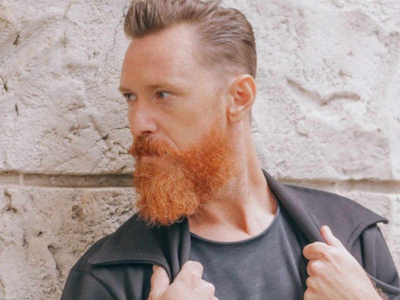 Ator blumenauense Imagem mostra um homem de cabelos e barbas ruivas posando para foto. Ele não olha para a câmera, apenas para o lado e segura um casaco da cor preta