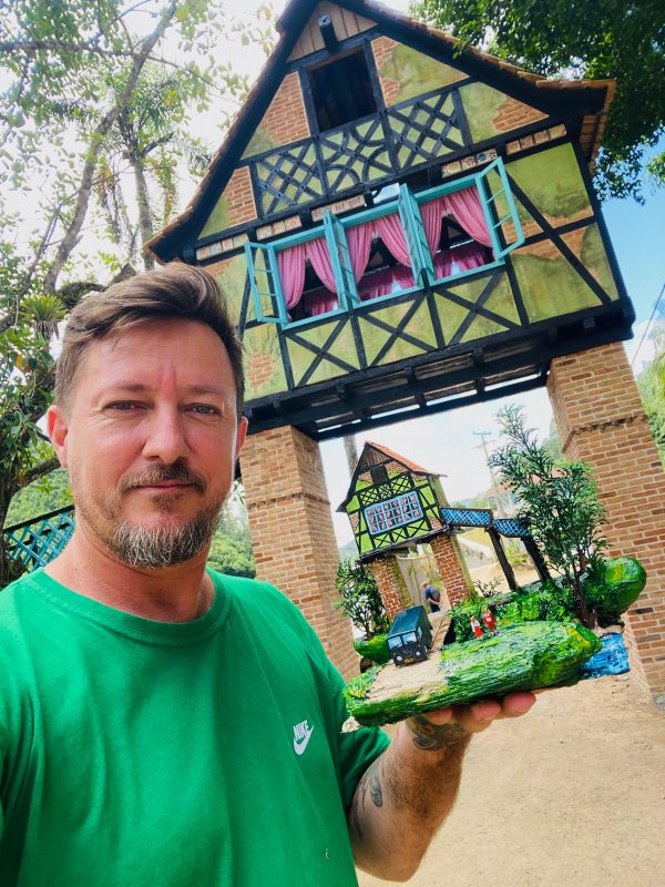 Imagem mostra morador de Blumenau que cria mini mundo germânico com materiais reciclados segurando uma miniatura em frente do portal que ele fez réplica