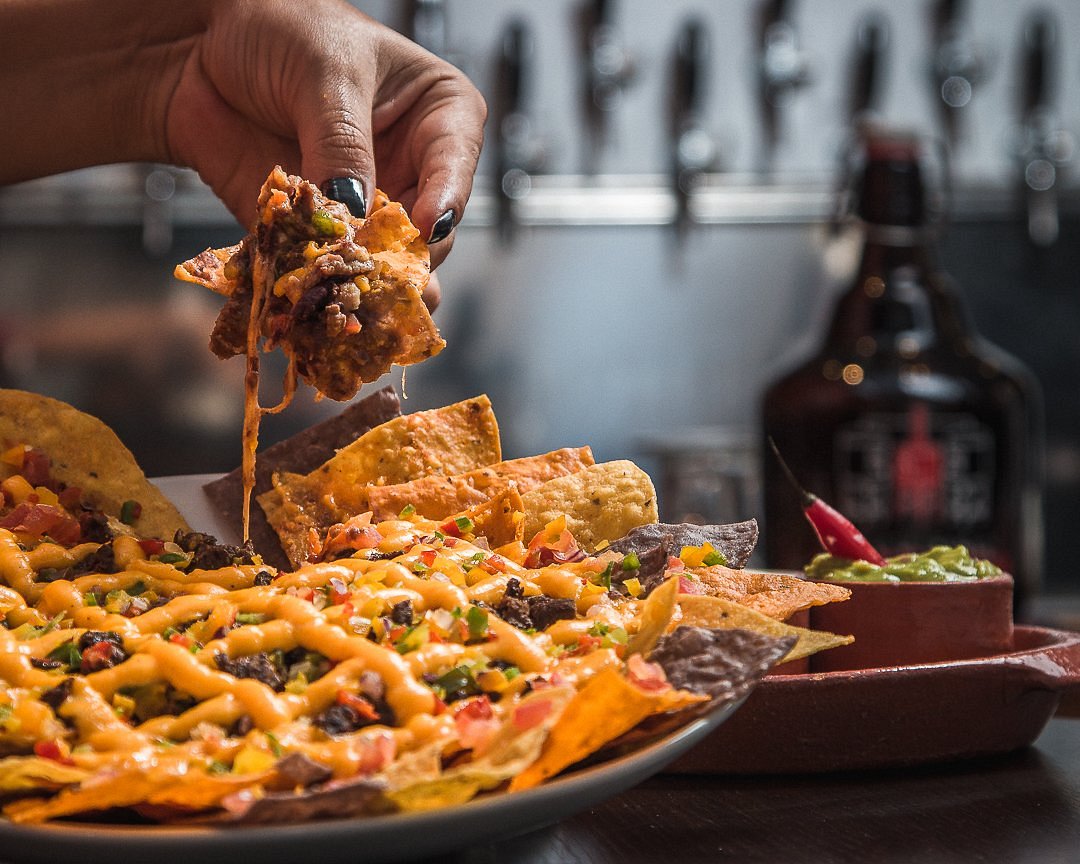 Foto de uma travessa com nachos, carne e queijo cheddar sobre uma mesa. Atrás da travessa, a mão de uma mulher segura um dos nachos no alto com carne e queijo escorrendo. O fundo é desfocado.
