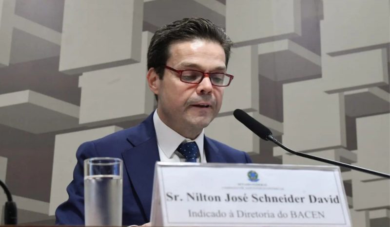 Nilton David alertou sobre possibilidade de recessão global, foto mostra homem de cabelos pretos, branco, com óculos de armação vermelha e terno azul falando em microfone