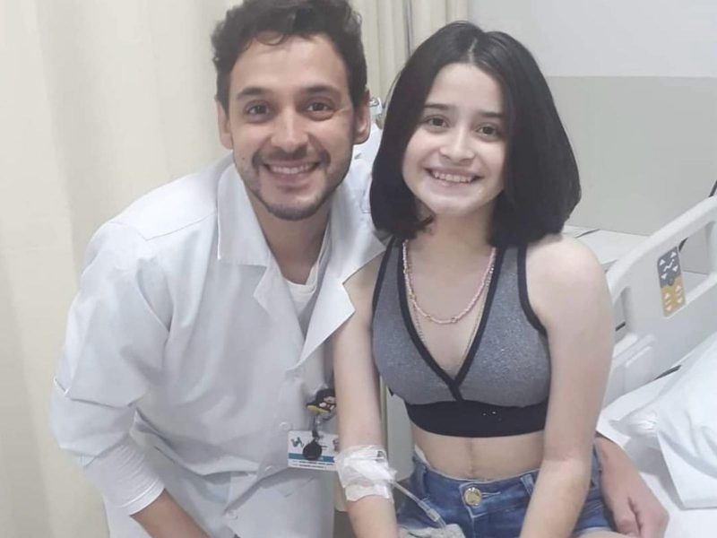 Imagem mostra um homem e uma jovem. O homem tem cabelo curto escuro, barba, está sorrindo para foto e usa jaleco de médico. A menina tem cabelos escuros, médios, também sorri para a foto, usa um top na cor cinza e um shorts jeans. No braço ela tem soro do hospital