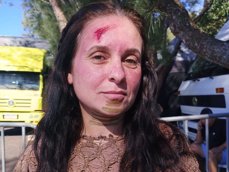 Atriz Blumenauense Imagem mostra uma mulher, de pele branca, com uma maquiagem que lembra machucados. Ela tem cabelo castanho escuro. Atrás dela tem uma árvore e um caminhão amarelo