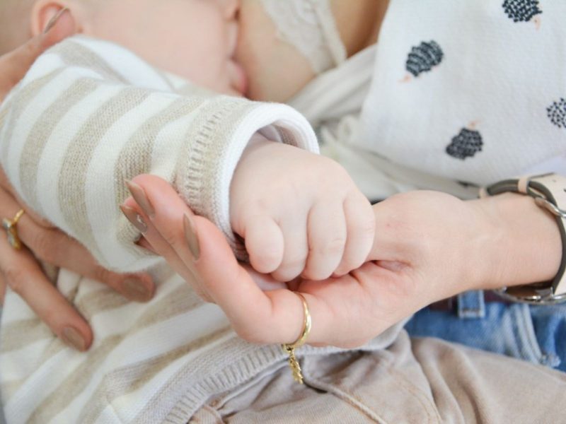 Amamentação Imagem mostra mãe amamentando bebê. Eles vestem roupas em tom bege claro e a mãe segura a mão do bebê, enquanto banco de Leite de Blumenau
