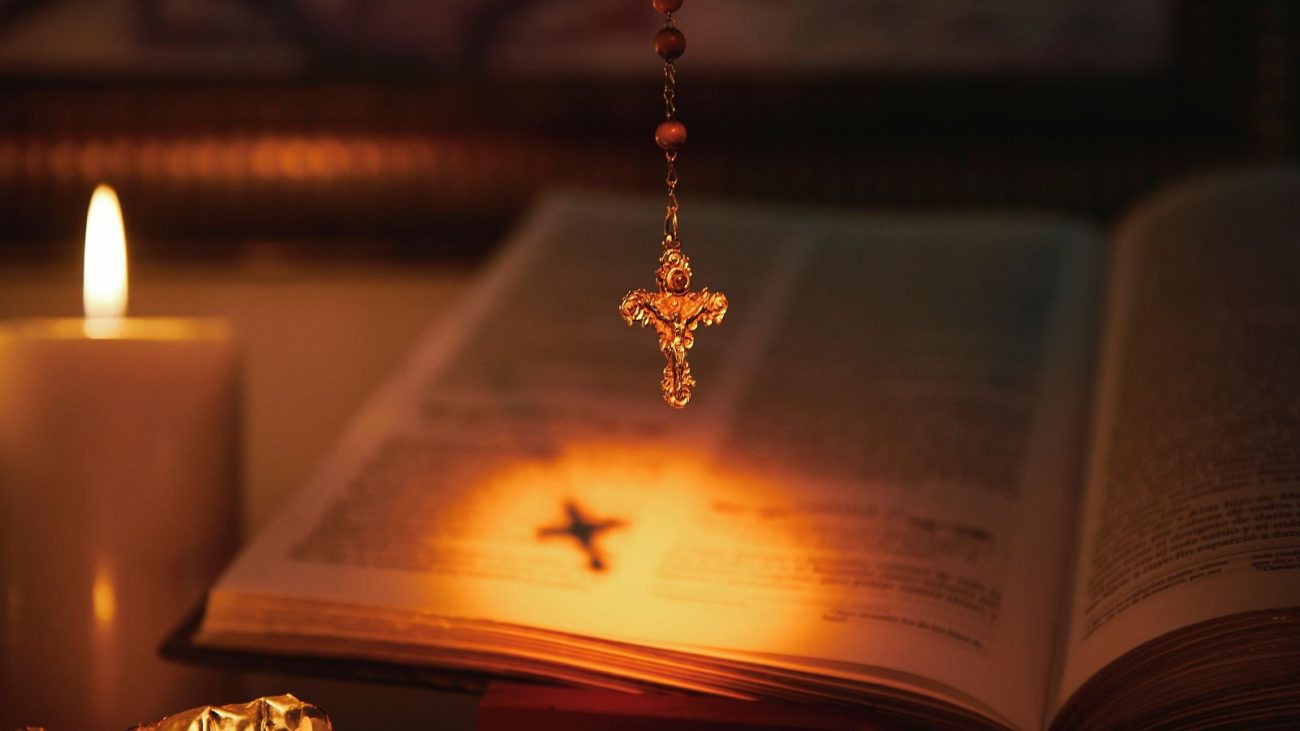 Santo do dia Imagem mostra um terço pendurado com a imagem de uma bíblia e uma vela acesa no fundo, para ilustrar o Santo do Dia em 28 de maio.