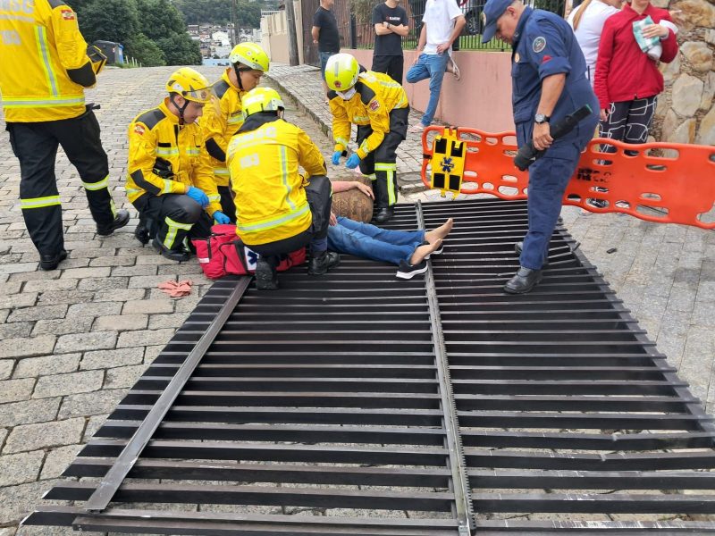 Acidente com portão Imagem mostra seis bombeiros socorrendo uma mulher que está desmaiada no chão. Ela está sob um portão na cor preta, que caiu após ser derrubado por um carro desgovernado