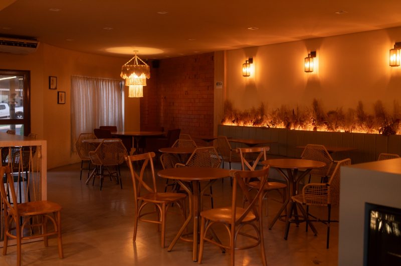 OCA Café Chapecó Ambiente interno do OCA Café em Chapecó. O chão é de lâminas de madeira, as mesas são pequenas, redondas e de madeira. O ambiente tem uma luz amarelada que o deixa mais escuro. Uma das paredes é de tijolos à vista e há algumas folhas secas numa grande floreira de concreto encostada em uma das paredes.