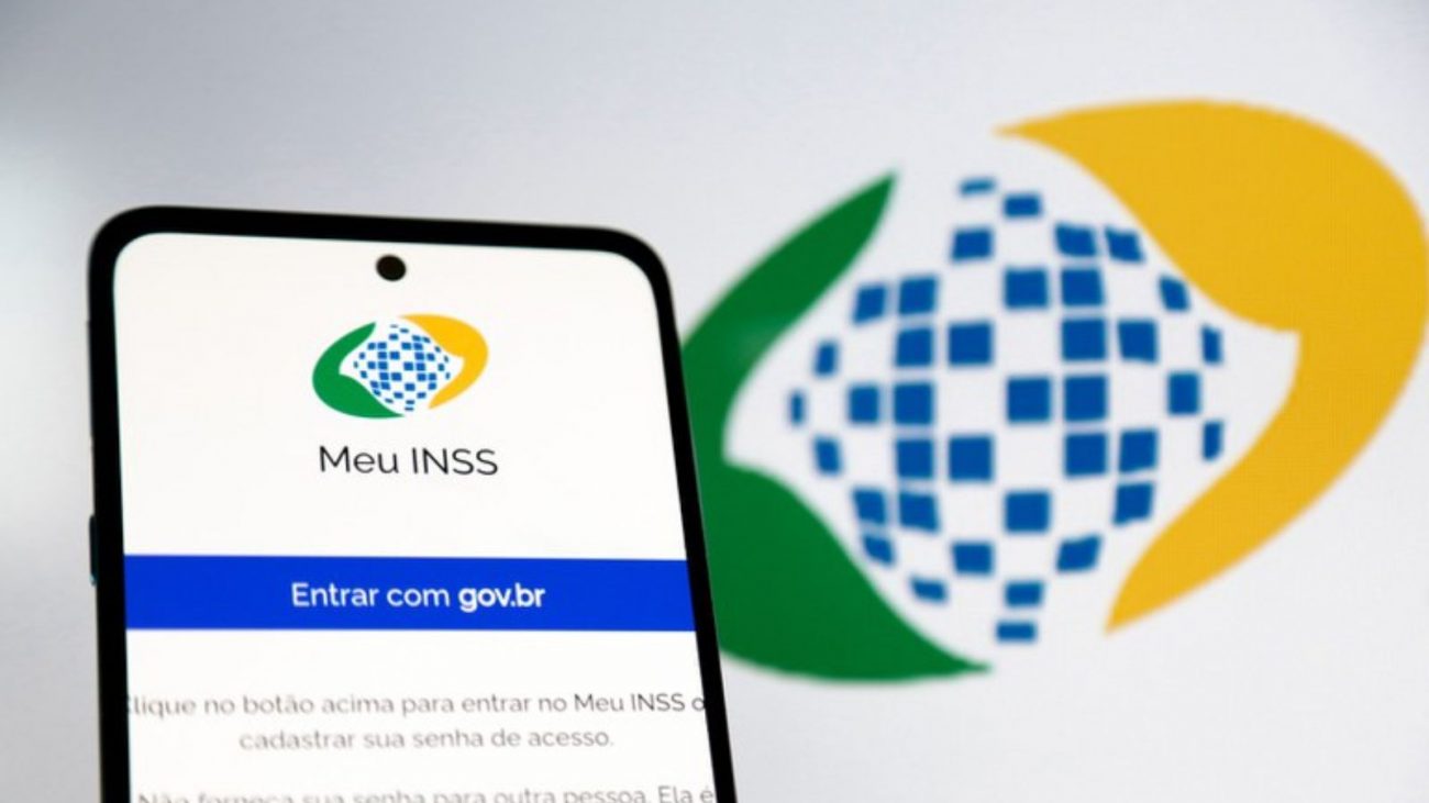 Imagem mostra o aplicativo Meu INSS em uma tela de celular com o símbolo do INSS ao fundo desfocado. A imagem serve para ilustrar a Calculadora do INSS, recurso disponível no Meu INSS.