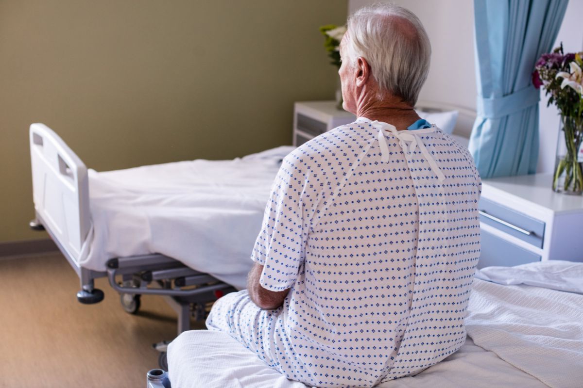 Homem idoso sentado na beira de uma cama de hospital, de costas para a câmera. Ele veste um avental hospitalar branco com pequenos desenhos azuis. Ao fundo, há uma cama hospitalar desocupada, uma mesa de cabeceira e um buquê de flores sobre um móvel, sugerindo um ambiente de internação ou recuperação médica