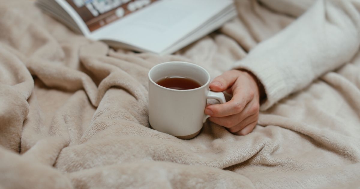 Imagem de uma pessoa na cama com cobertor e caneca, para indicar que a previsão do tempo terá temperaturas mais agradáveis 