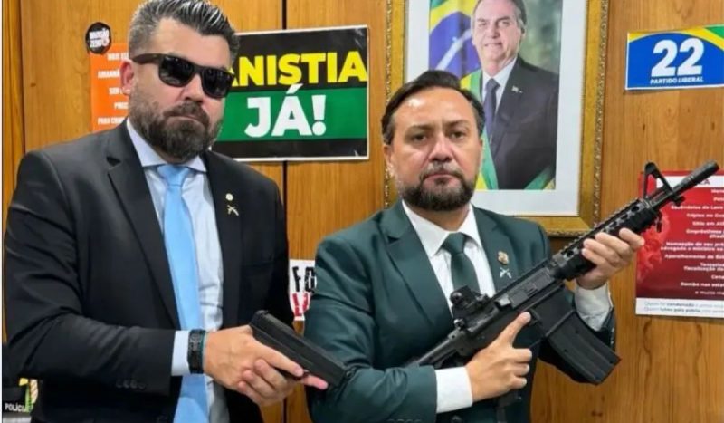 Deputado federal Delegado Caveira segurando uma pistola ao lado do vereador Zezinho Lima Delegado Caveira, o deputado que levou fuzil para a Câmara publicou foto em seu gabinete segurando uma pistola ao lado do vereador Zezinho Lima, que segura um fuzil
