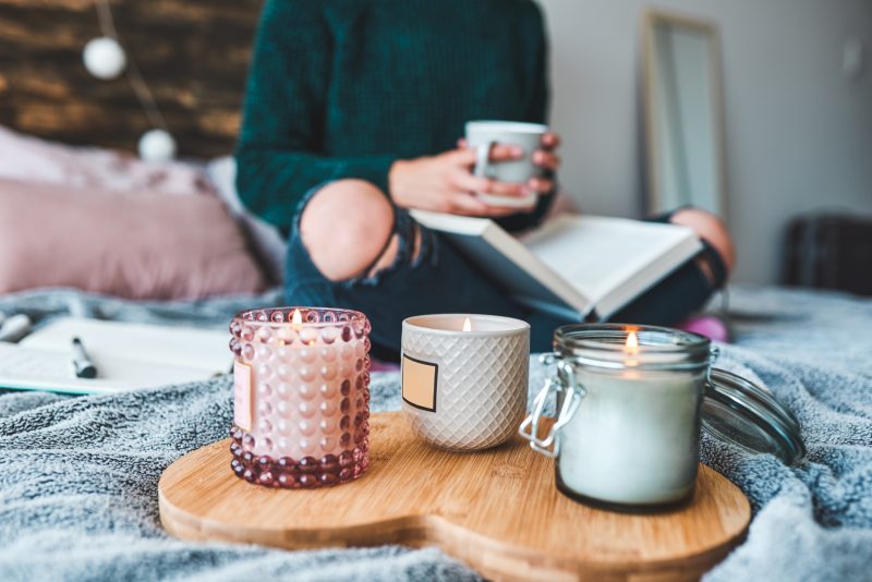 Mulher lendo um livro e segurando uma xícara de chá, com velas aromáticas na cama para deixar a casa cheirosa antes de dormir