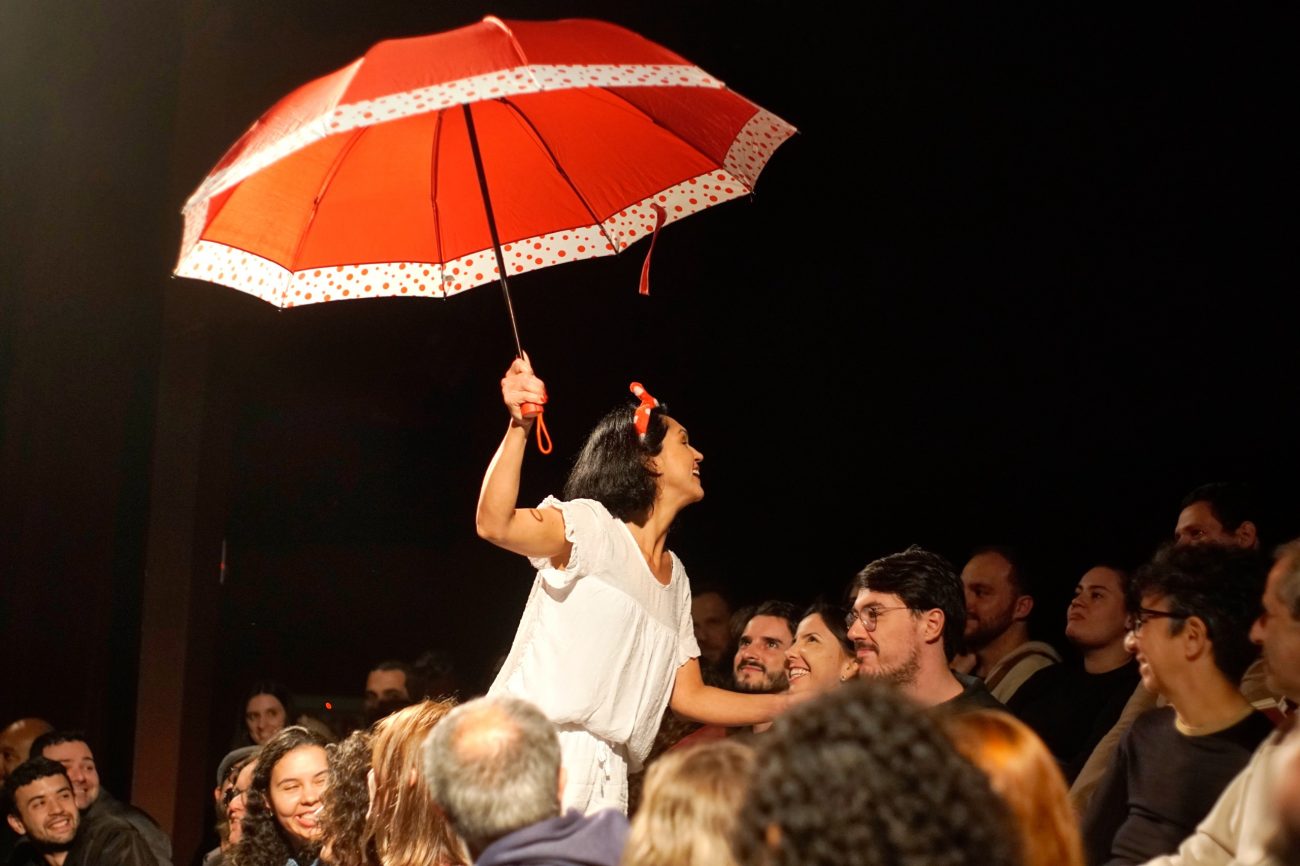Festival Nova Cena Mulher segura um guarda-chuva vermelho enquanto atua no Festival Nova Cena