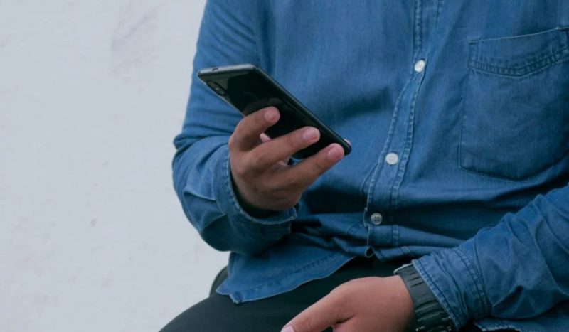 Homem de camisa azul mexendo no celular Funcionário furtou R$ 13 mil do patrão ao registrar ponto pelo celular da empresa