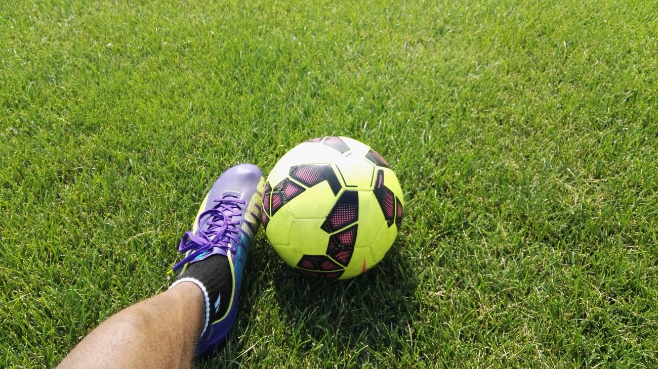 Imagem mostra a perna de um jogador de futebol ao lado de uma bola em campo. Artigo sobre handicap