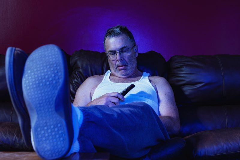 Sedentarismo Homem de meia-idade sedentário assistindo TV no sofá para representar hábitos prejudiciais para a saúde após os 50 anos
