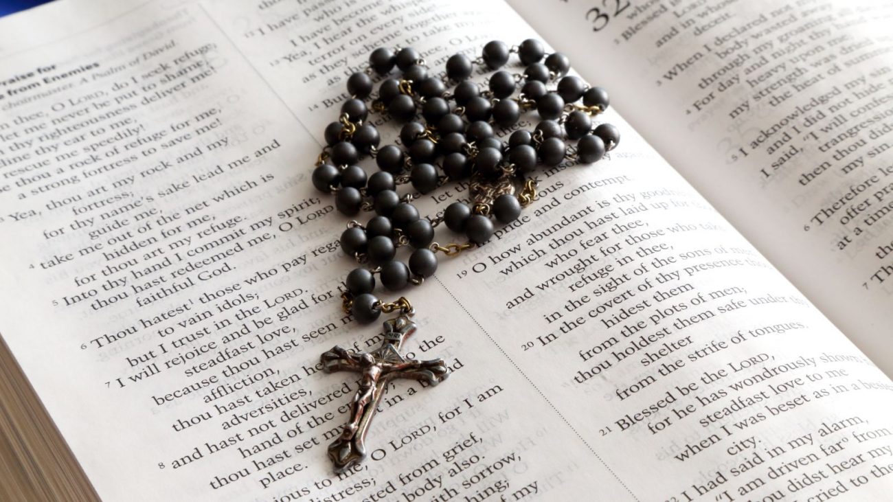Imagem mostra uma bíblia com um terço preto em cima para simbolizar o santo do dia. 