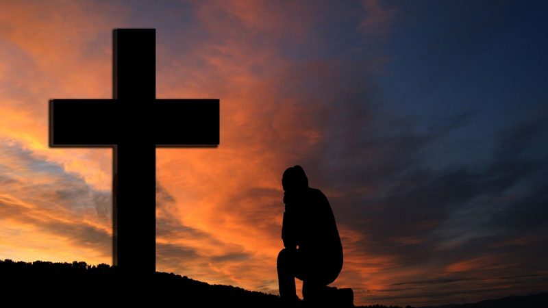 Imagem de uma cruz contra luz e uma pessoa à frente rezando para simbolizar São Cristóvão. 